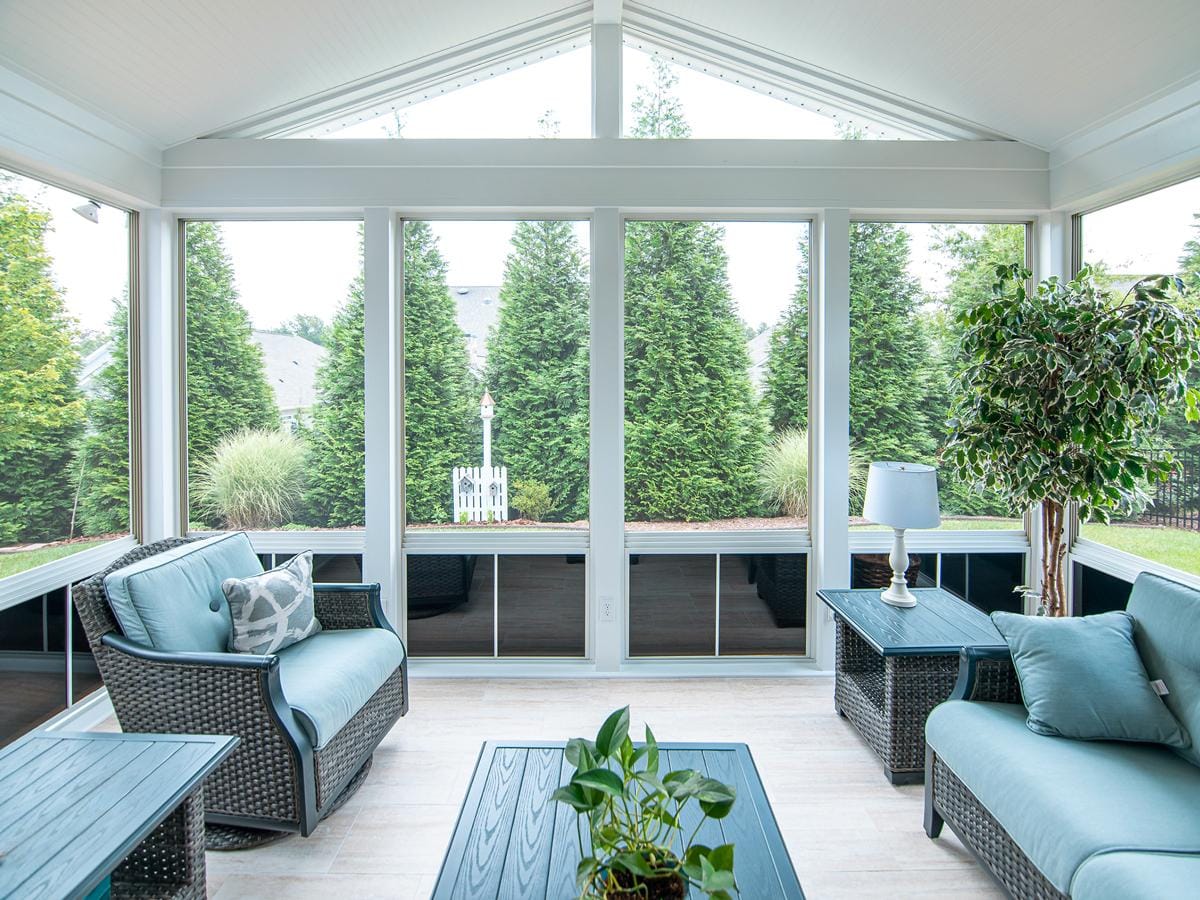 Interior of a three-season room with a vaulted ceiling, glass walls, and wicker seating, looking out to a tree-lined South Jersey yard